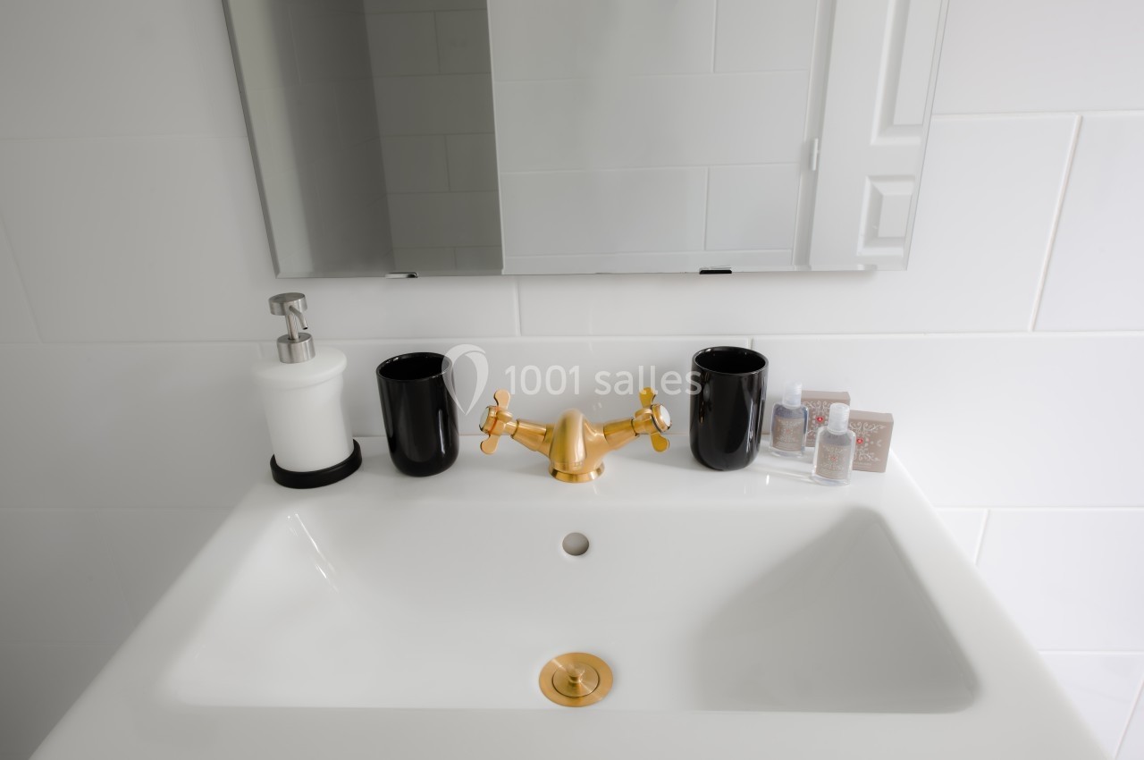 Lavabo blanc avec robinet doré, deux gobelets noirs, un distributeur de savon et un miroir mural.