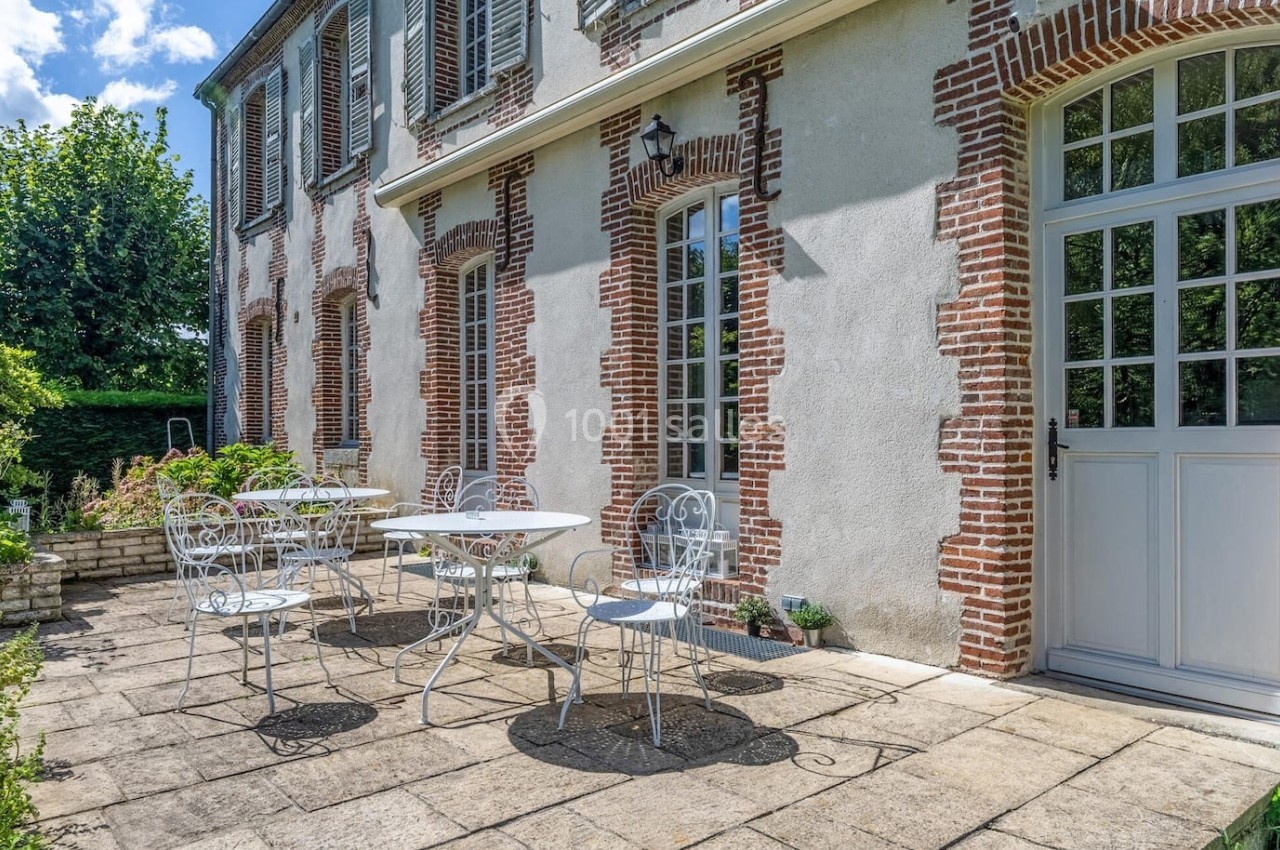 Terrasse en pierre avec tables et chaises en fer forgé devant une maison en briques et crépi, entourée de verdure.
