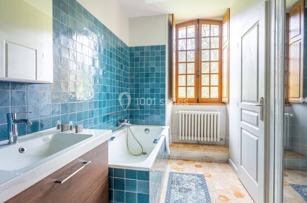 Salle de bain lumineuse avec carrelage bleu, baignoire, lavabo moderne et fenêtre en bois donnant sur l'extérieur.