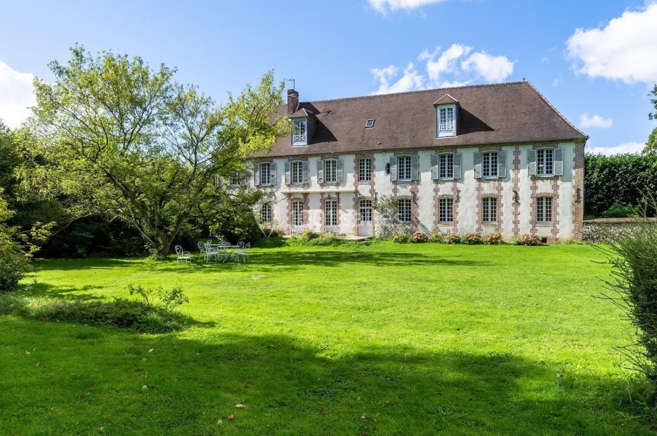 Grande maison en pierre avec toit en ardoise, entourée d'un vaste jardin verdoyant sous un ciel bleu.