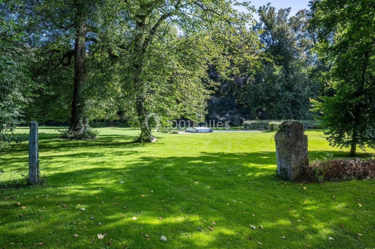 Pelouse verdoyante entourée d'arbres, avec une pierre tombale et un monument en arrière-plan sous un ciel dégagé.