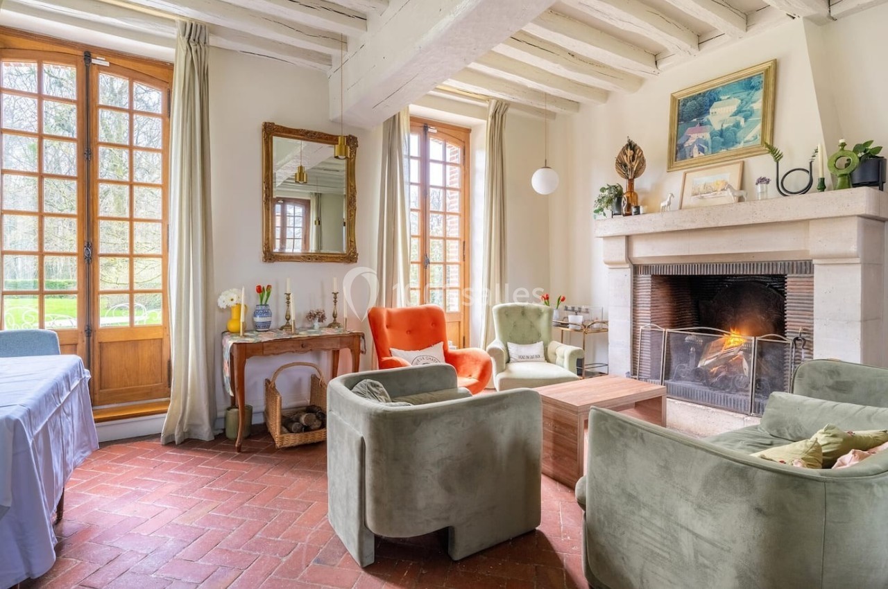 Salon lumineux avec cheminée allumée, fauteuils confortables, grandes fenêtres et poutres apparentes.