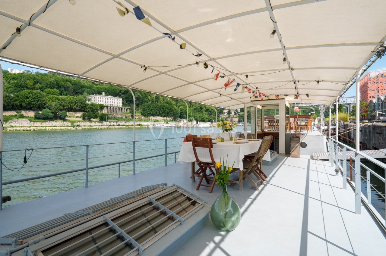 Terrasse couverte d'un bateau aménagée avec des tables dressées, chaises en bois et vue sur une rivière bordée de verdure.