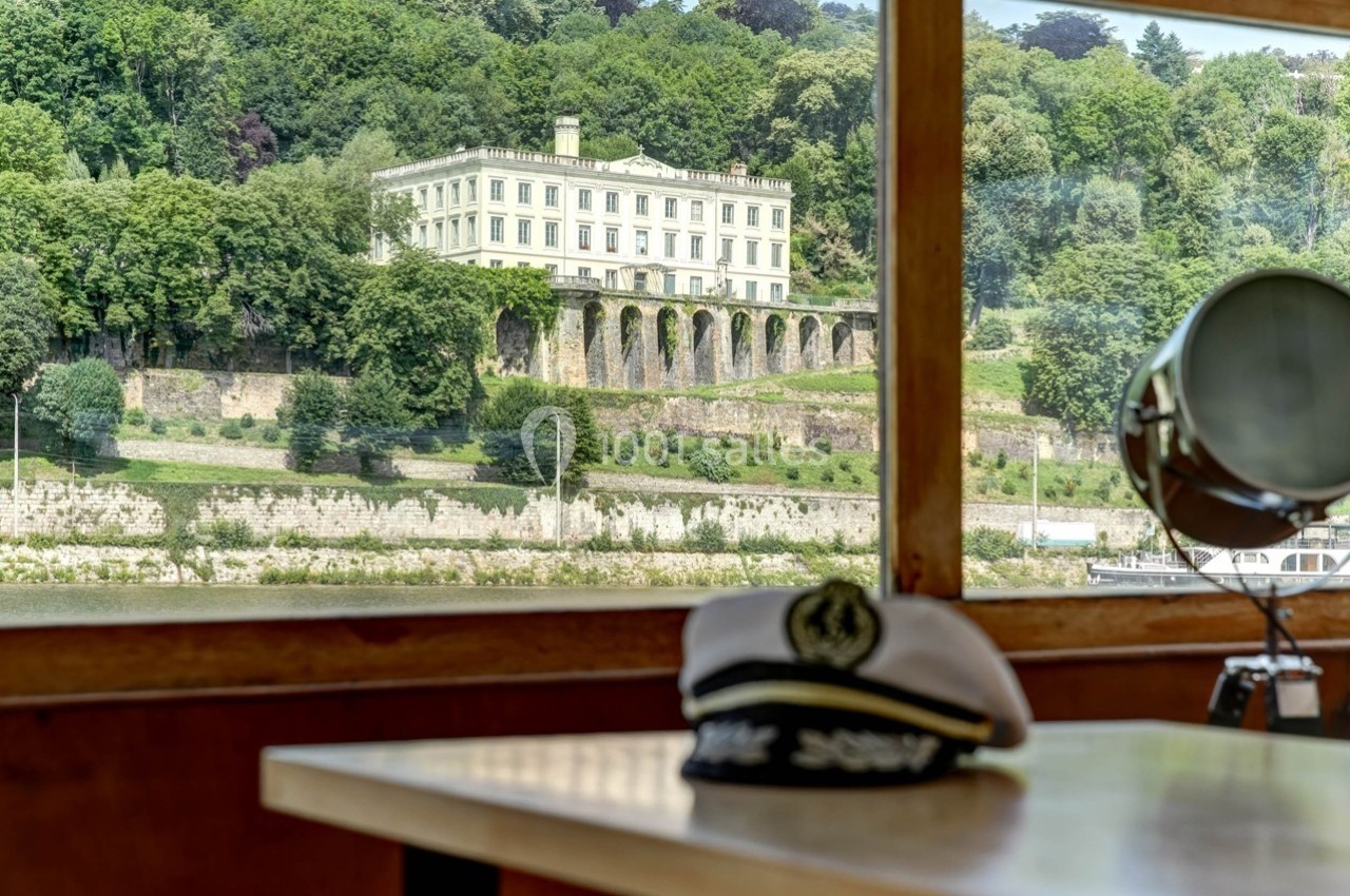 Vue d'un bâtiment historique entouré de verdure depuis l'intérieur d'un bateau, avec une casquette de marin au premier plan.