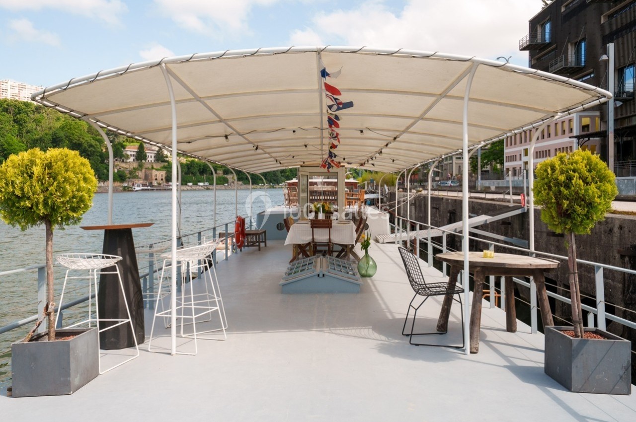 Terrasse d’un bateau aménagée avec des tables, des chaises et des plantes, amarré près d’un quai urbain.