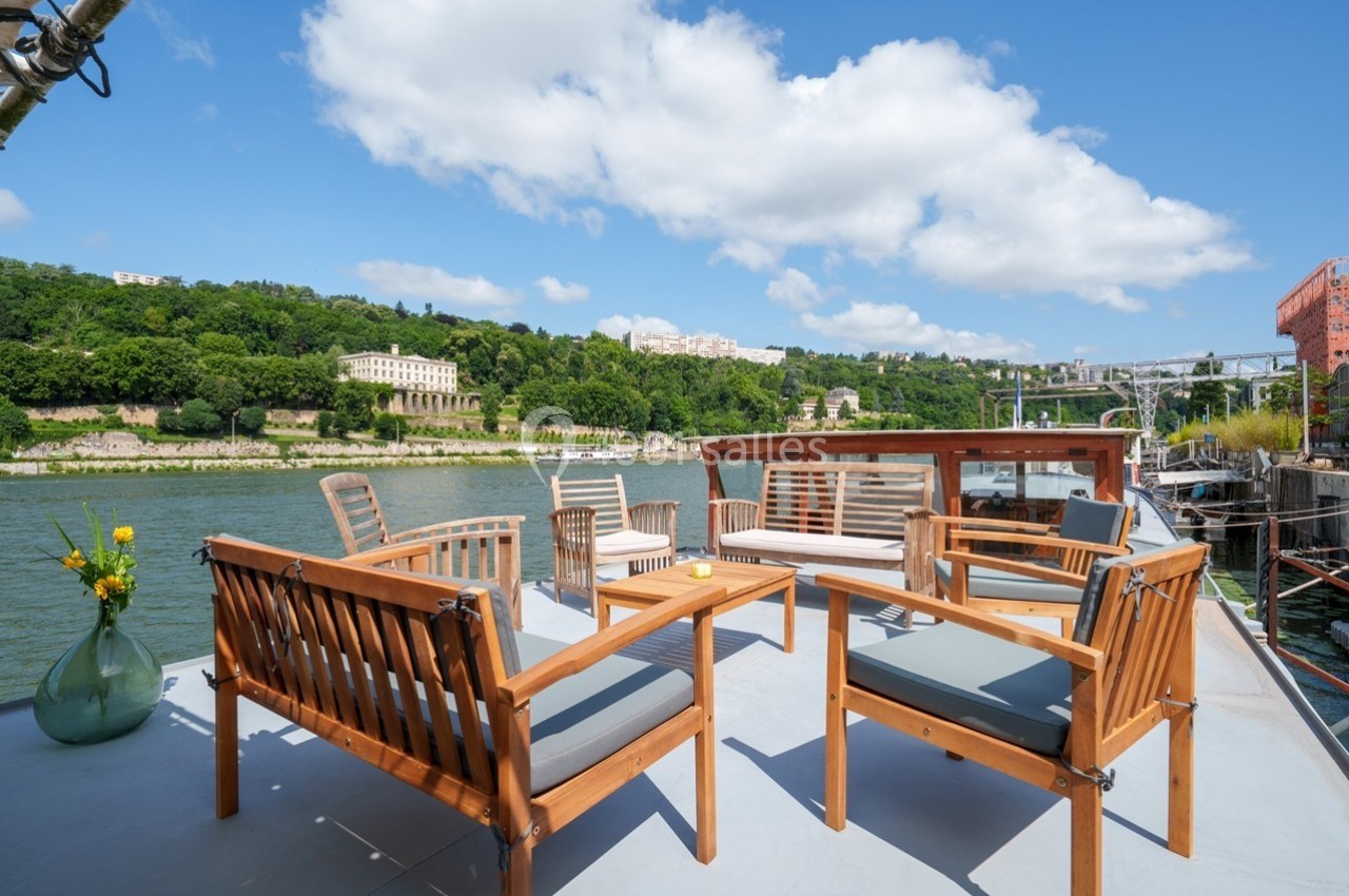 Terrasse en bois aménagée avec des fauteuils, offrant une vue sur une rivière et des collines verdoyantes sous un ciel bleu.
