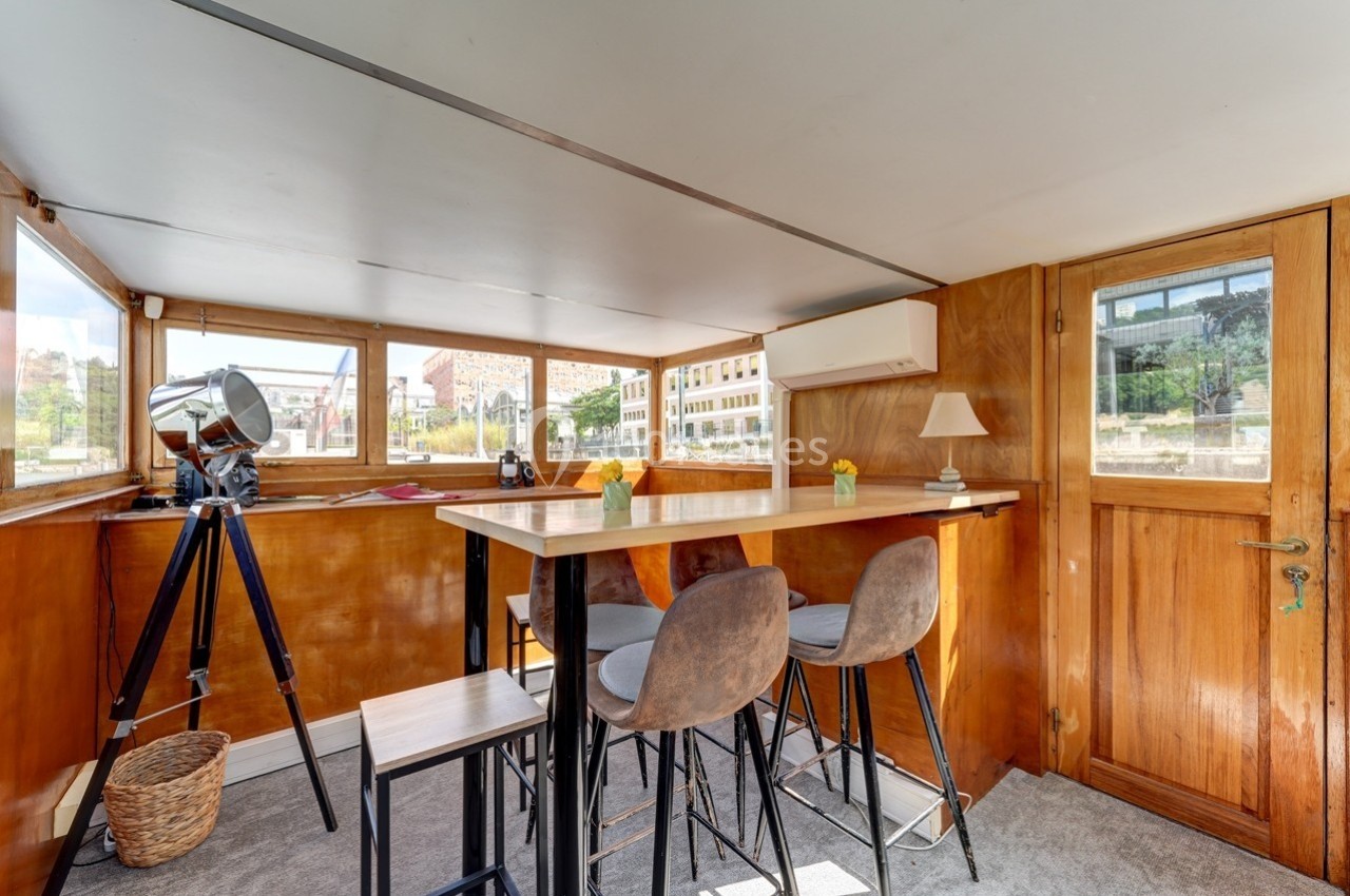 Intérieur lumineux d'une péniche aménagée avec table haute, tabourets, télescope et vue sur des bâtiments extérieurs.