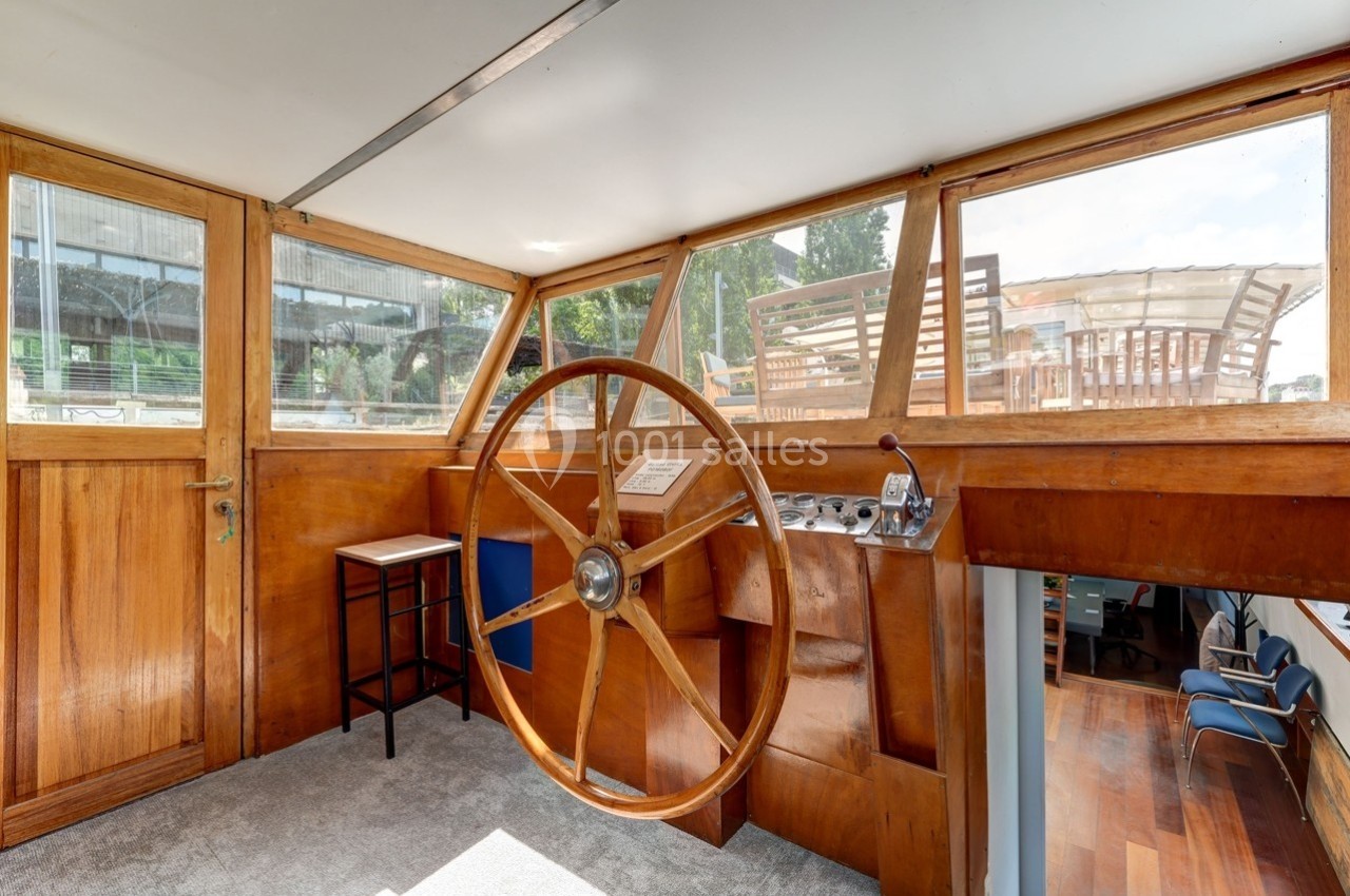Poste de pilotage en bois d'un bateau avec un grand gouvernail, entouré de fenêtres offrant une vue extérieure.
