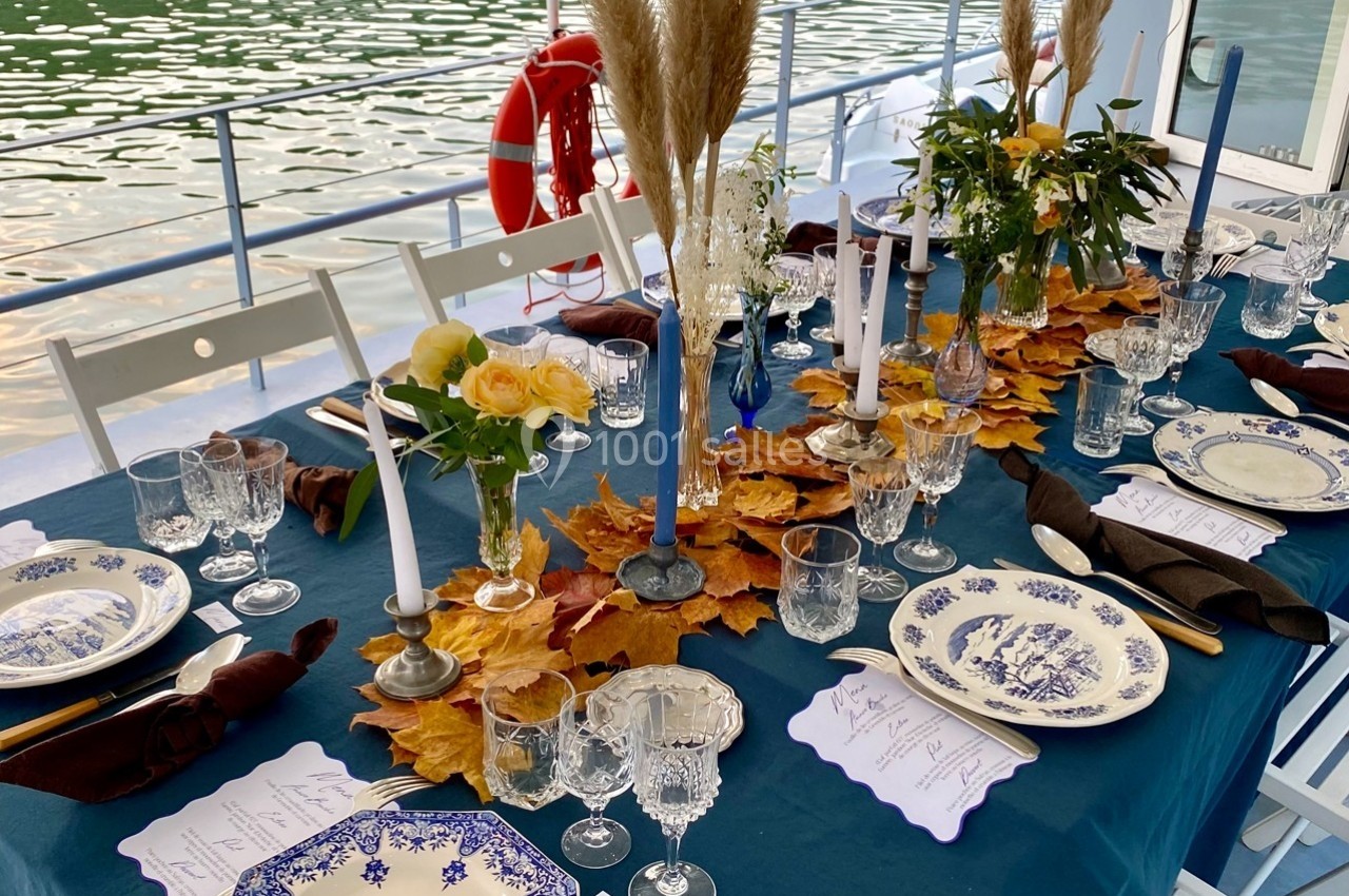 Table élégamment dressée sur une péniche, décorée de feuilles d'automne, bougies et vaisselle en porcelaine bleue.