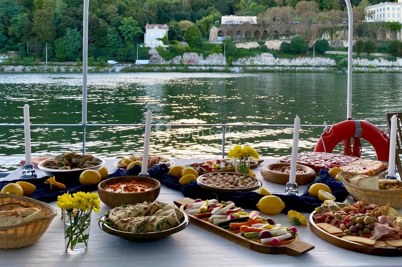 Table dressée avec des plats variés, bougies et fleurs, en extérieur près d'une rivière bordée de végétation.
