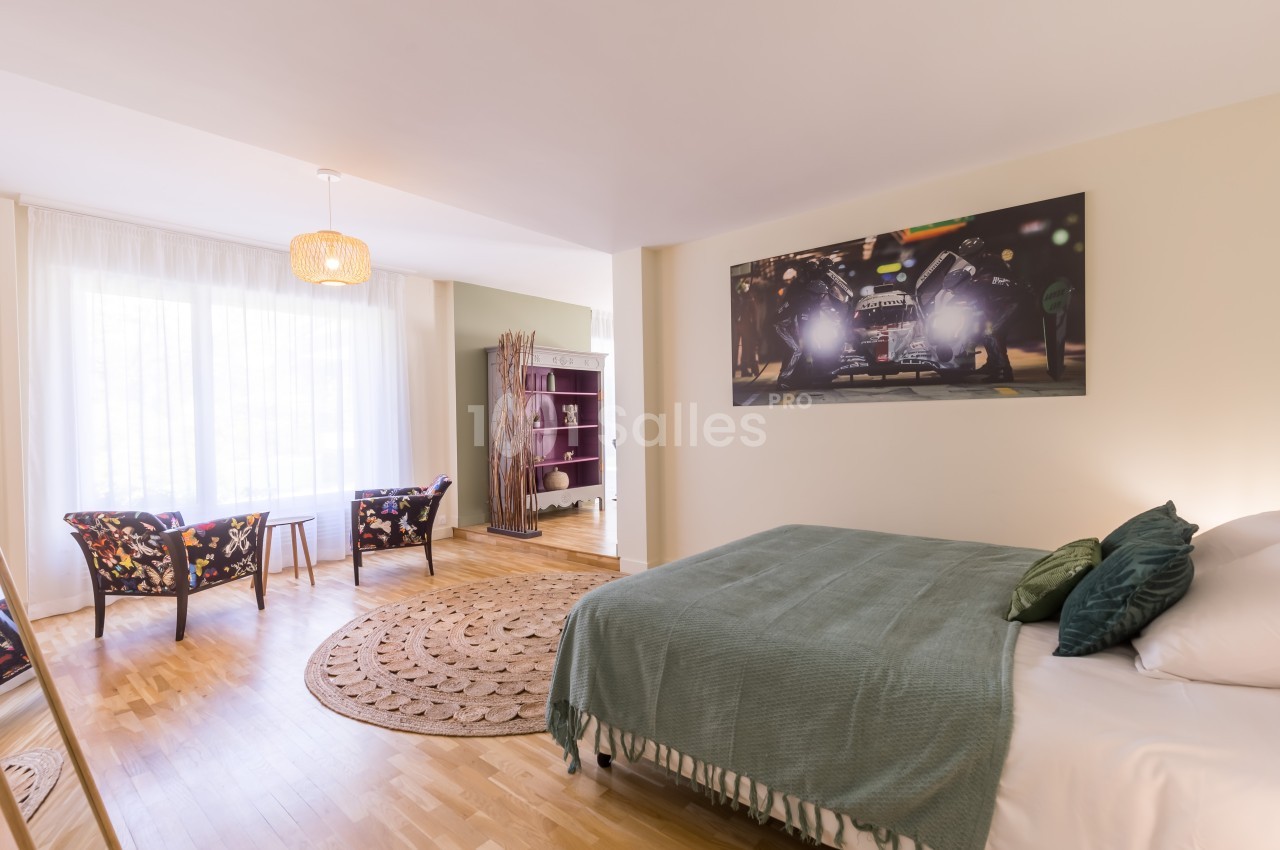 Chambre lumineuse avec un lit double, fauteuils près d'une grande fenêtre, tapis rond et étagère en arrière-plan.