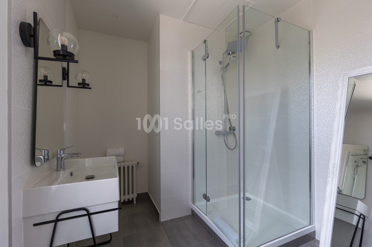 Salle de bain moderne avec douche vitrée, lavabo avec miroir et éclairage mural, murs blancs et sol gris.