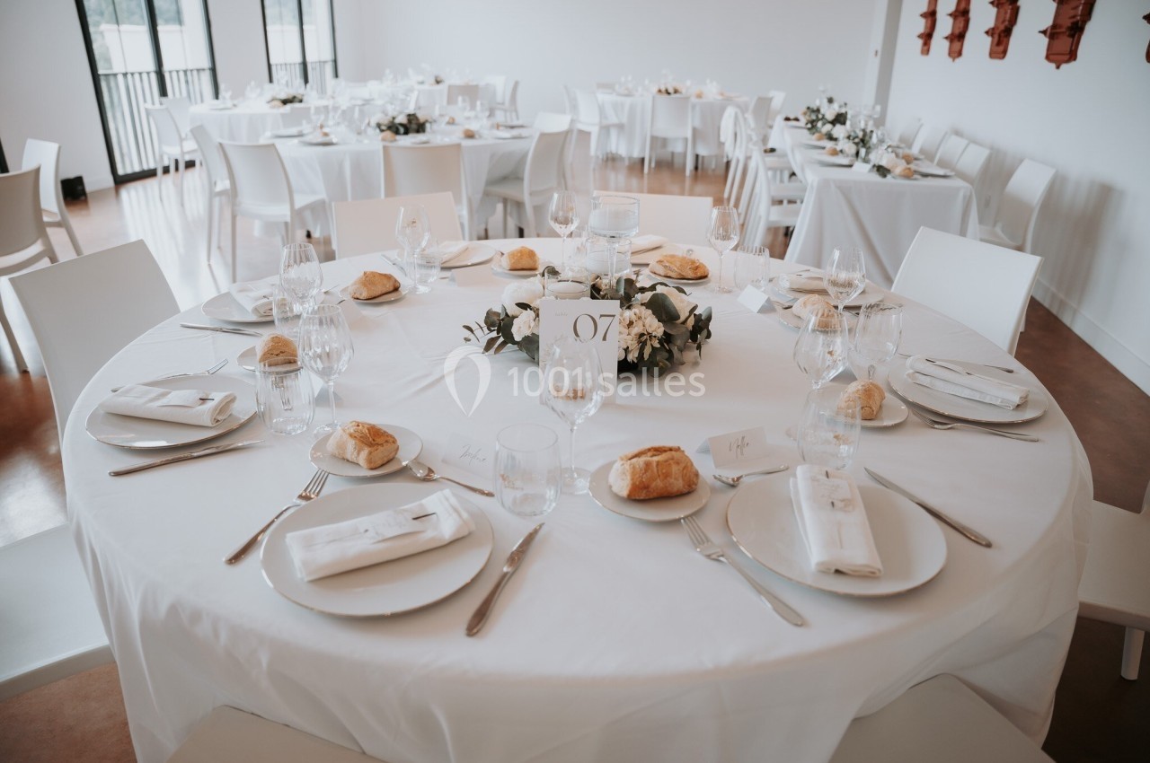 Table ronde élégamment dressée avec nappes blanches, couverts, assiettes, verres et décorations florales au centre.