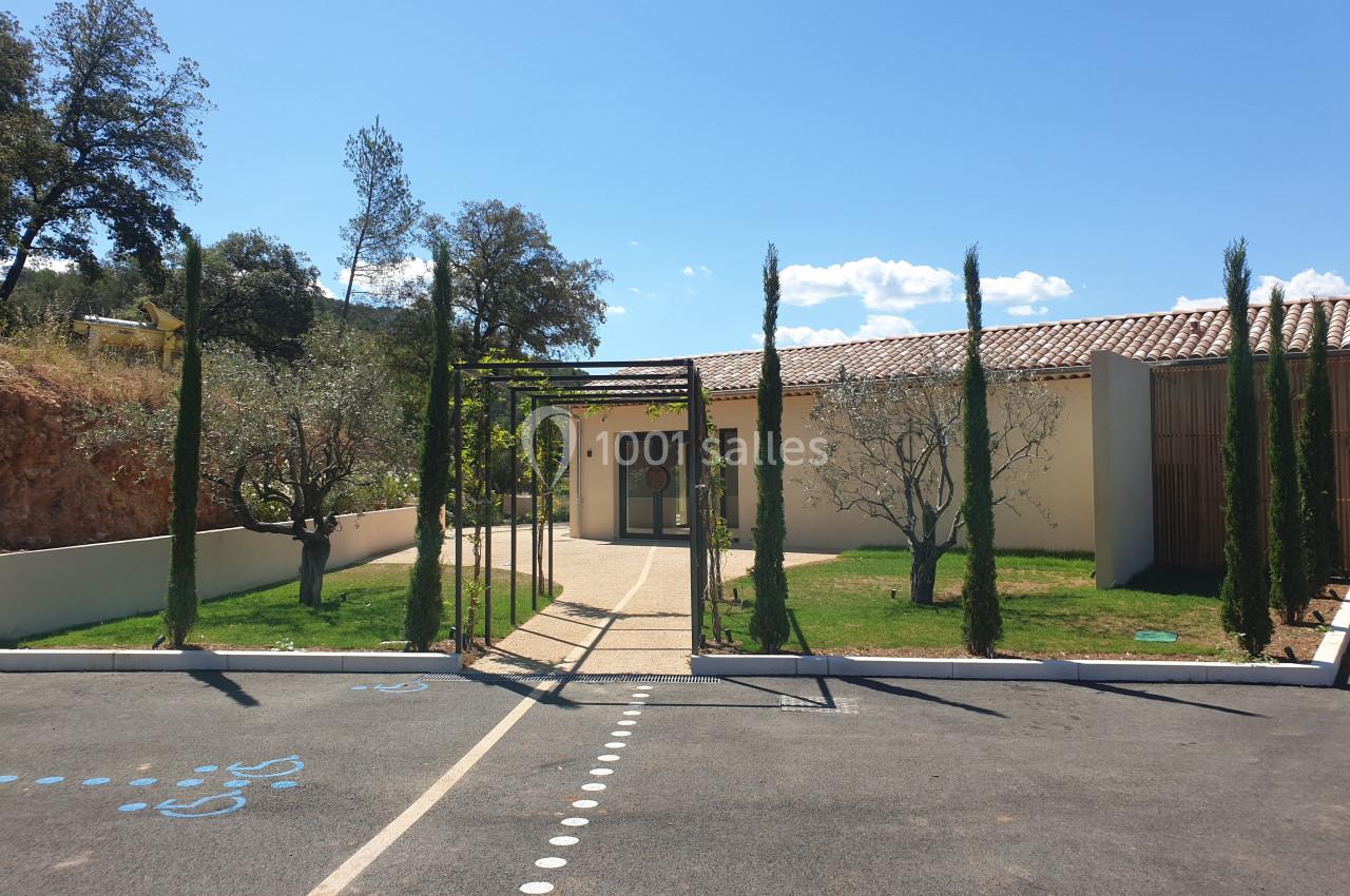 Entrée d'un bâtiment moderne entourée de cyprès et d'oliviers, avec un chemin menant à une porte vitrée.