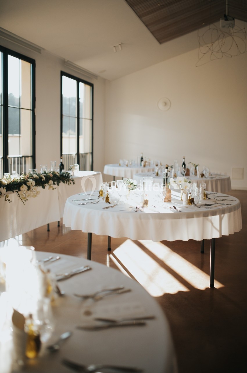 Salle lumineuse avec tables rondes dressées pour un repas, décorées de nappes blanches et de vaisselle élégante.