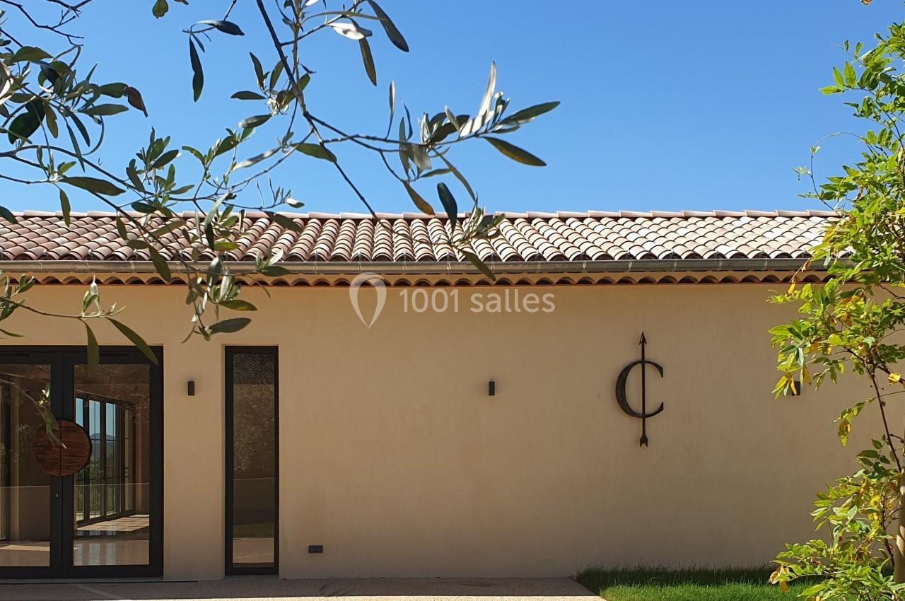 Façade d'une maison moderne avec toit en tuiles, porte vitrée, et végétation au premier plan sous un ciel bleu.