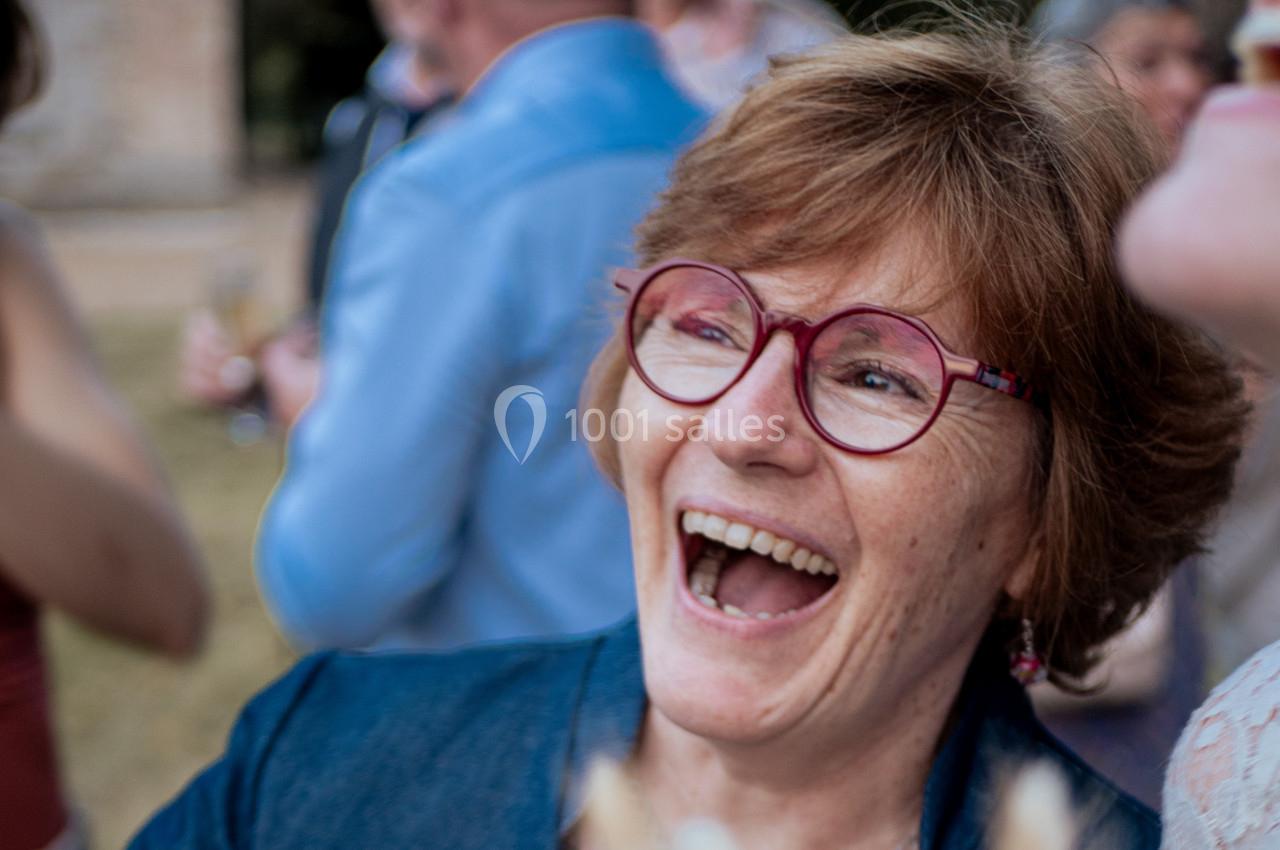 Femme souriante portant des lunettes rouges, entourée de personnes floues en arrière-plan lors d'un événement en extérieur.