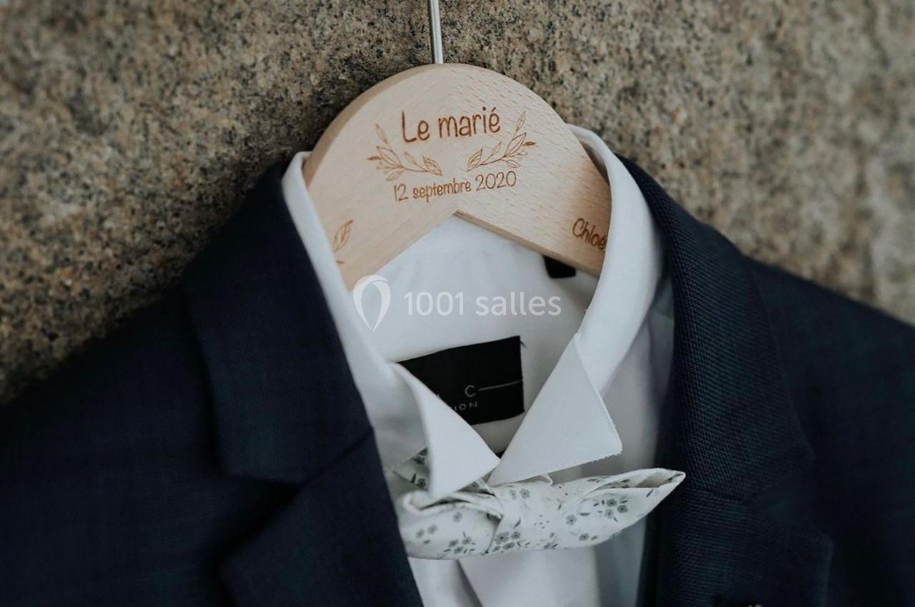 Costume de mariage bleu foncé avec chemise blanche et nœud papillon floral, suspendu sur un cintre gravé.