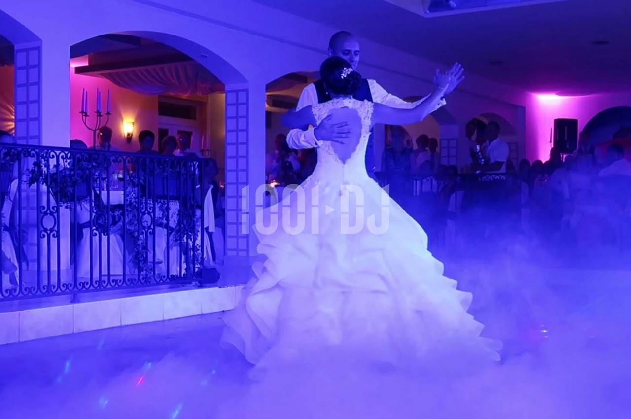 Un couple danse dans une salle éclairée par des lumières colorées, entouré de fumée au sol et d'invités.