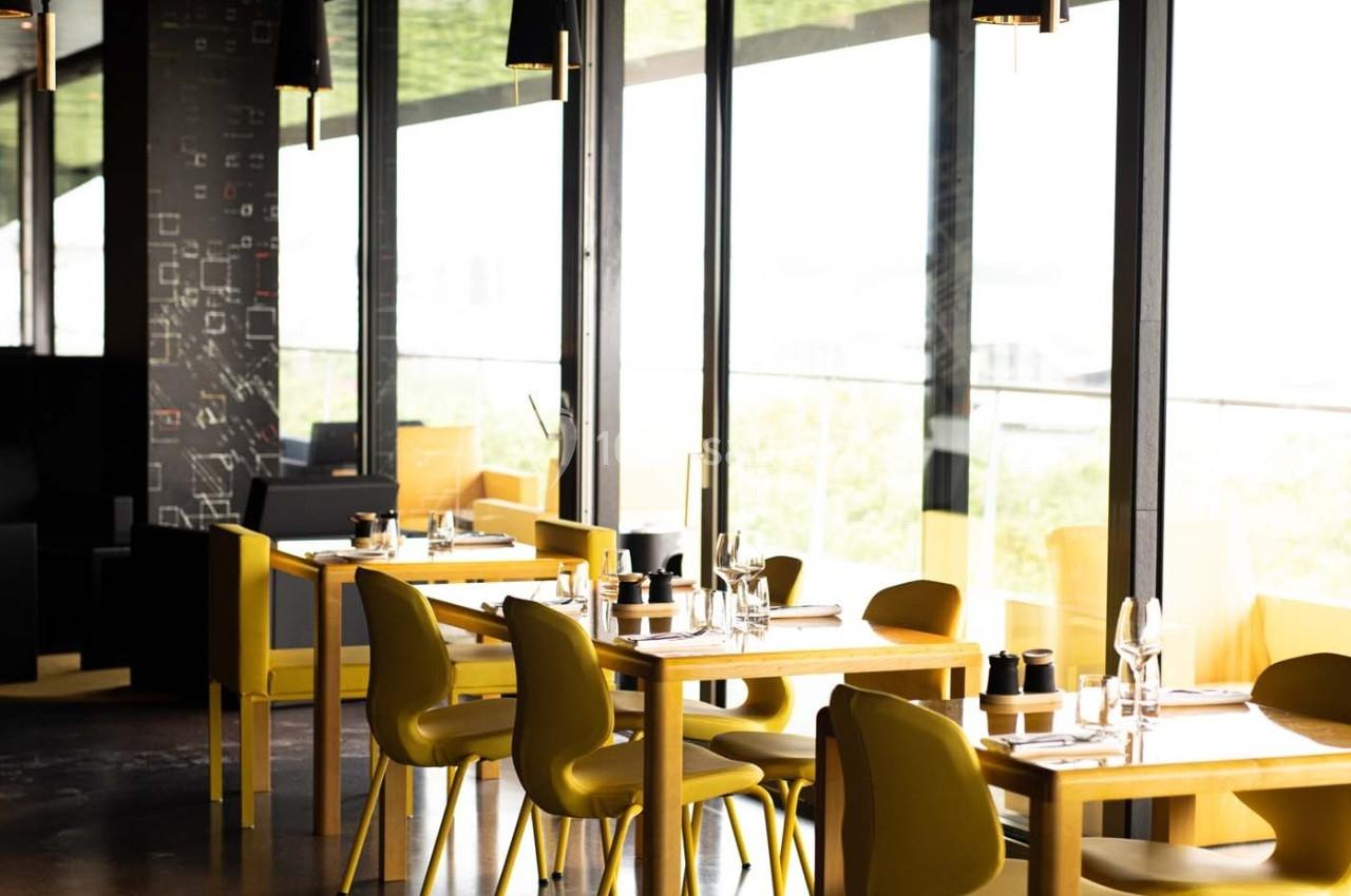 Salle de restaurant moderne avec des tables en bois clair, chaises jaunes et grandes baies vitrées laissant entrer la…