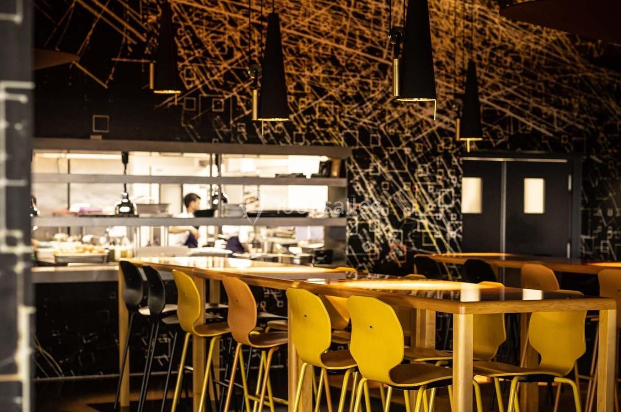Salle de restaurant moderne avec des tables en bois, chaises jaunes et vue sur une cuisine ouverte en arrière-plan.