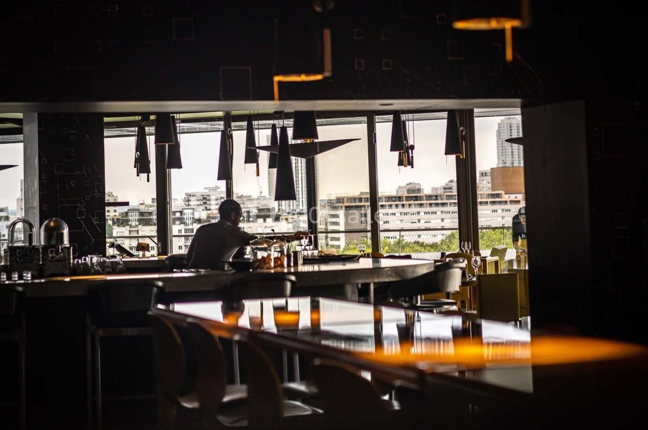 Un homme assis seul dans un bar sombre avec vue sur des immeubles à travers de grandes baies vitrées.
