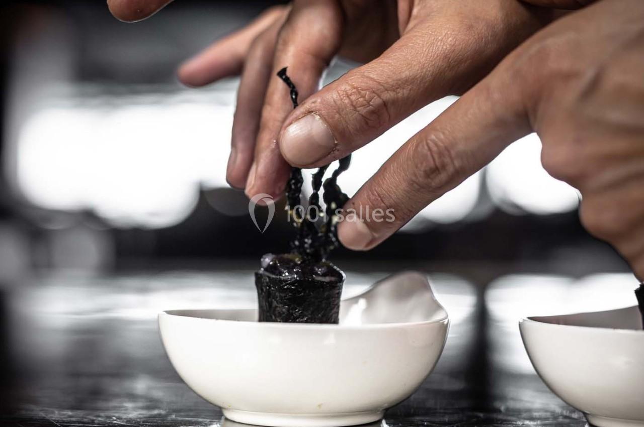 Deux mains assemblent un petit rouleau d'algues noires dans une coupelle blanche sur une surface sombre.