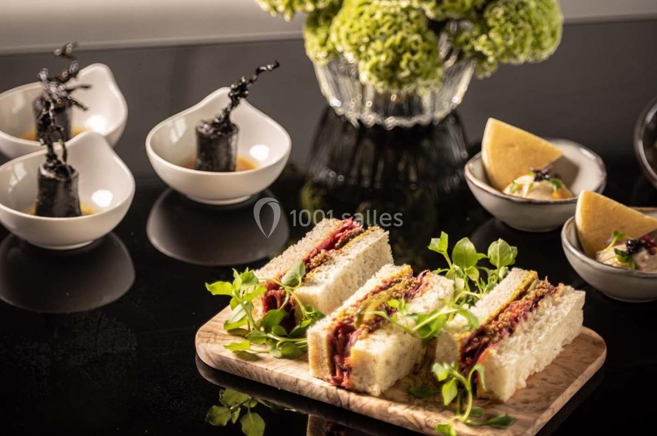 Assiette de sandwichs garnis de viande, accompagnée de petits plats et d'une décoration florale en arrière-plan.