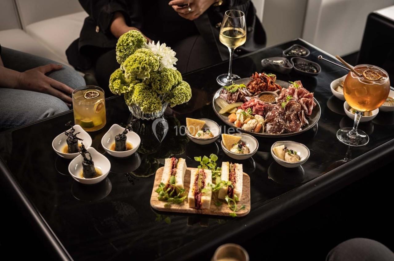 Table avec des amuse-bouches variés, cocktails et fleurs décoratives, entourée de convives en arrière-plan.