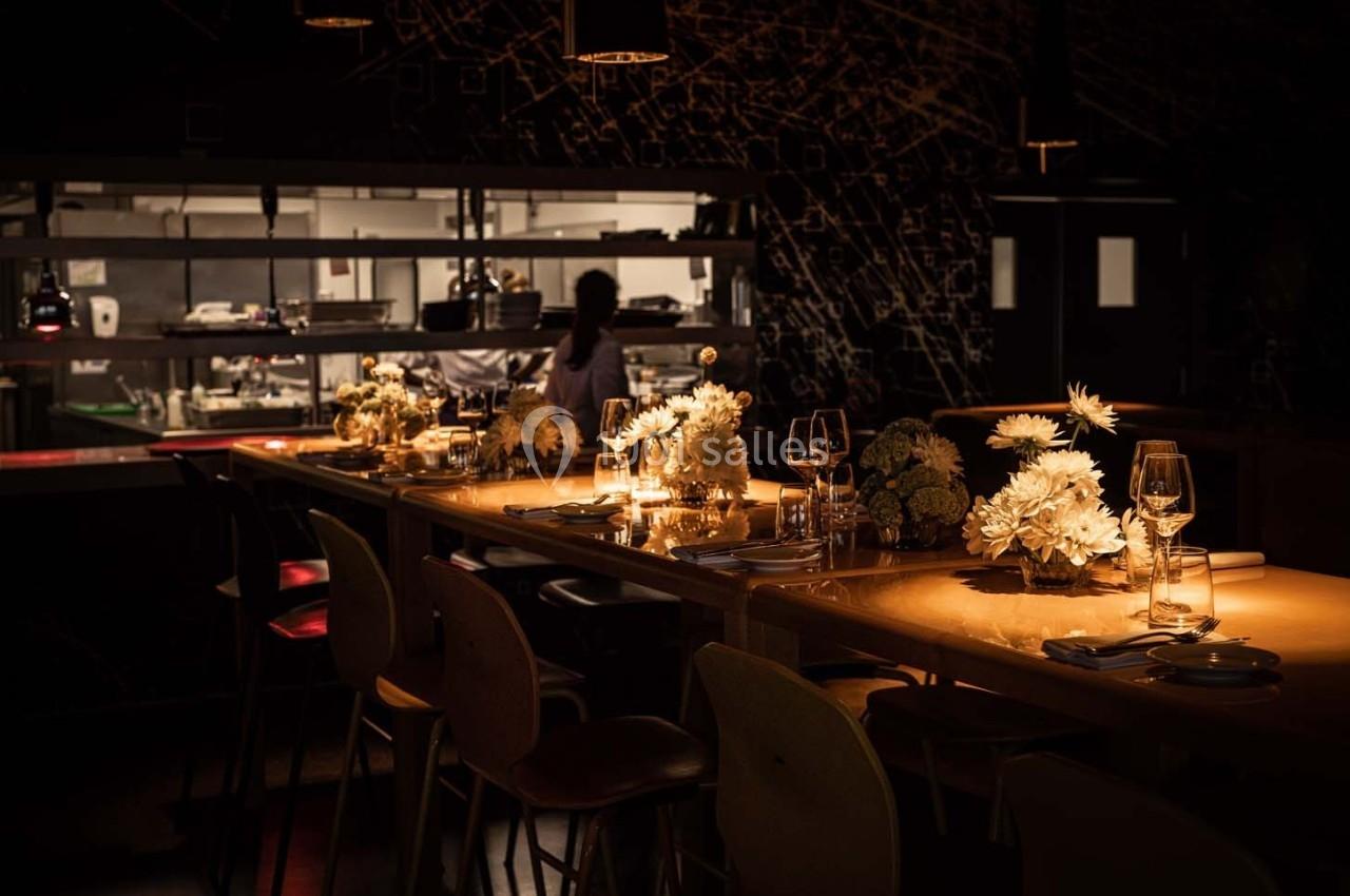 Salle de restaurant tamisée avec une grande table décorée de fleurs blanches, vue sur une cuisine ouverte en arrière-plan.