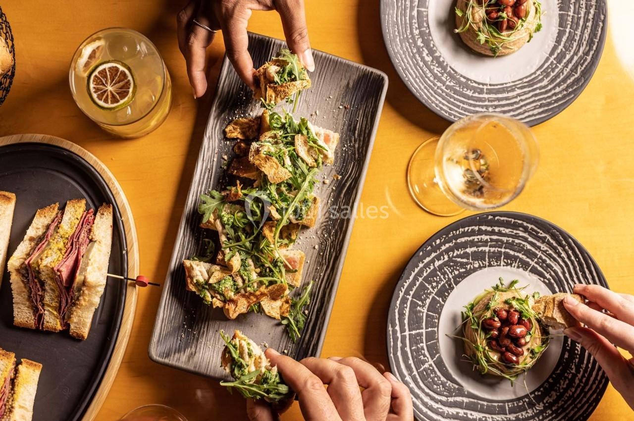 Assiette de toasts garnis de roquette et légumes, entourée de mains partageant le repas sur une table dressée.