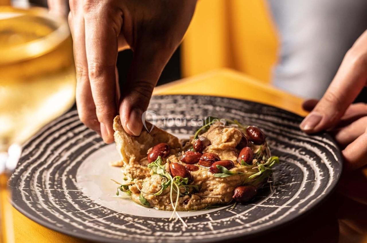 Une main trempe un morceau de pain dans un houmous garni de graines et de pousses sur une assiette noire.