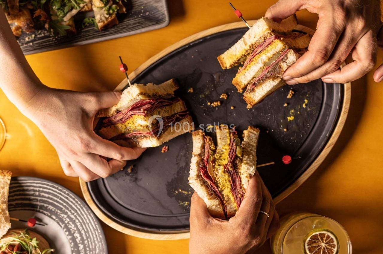 Trois mains tenant des sandwichs garnis de viande et de fromage sur un plateau en bois, avec des plats en arrière-plan.