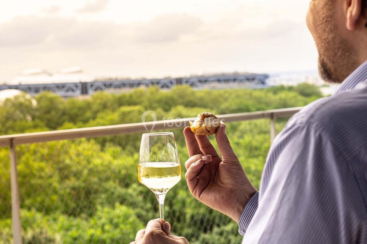 Un homme tient un verre de vin blanc et un amuse-bouche sur une terrasse avec vue sur des arbres et un bâtiment au loin.