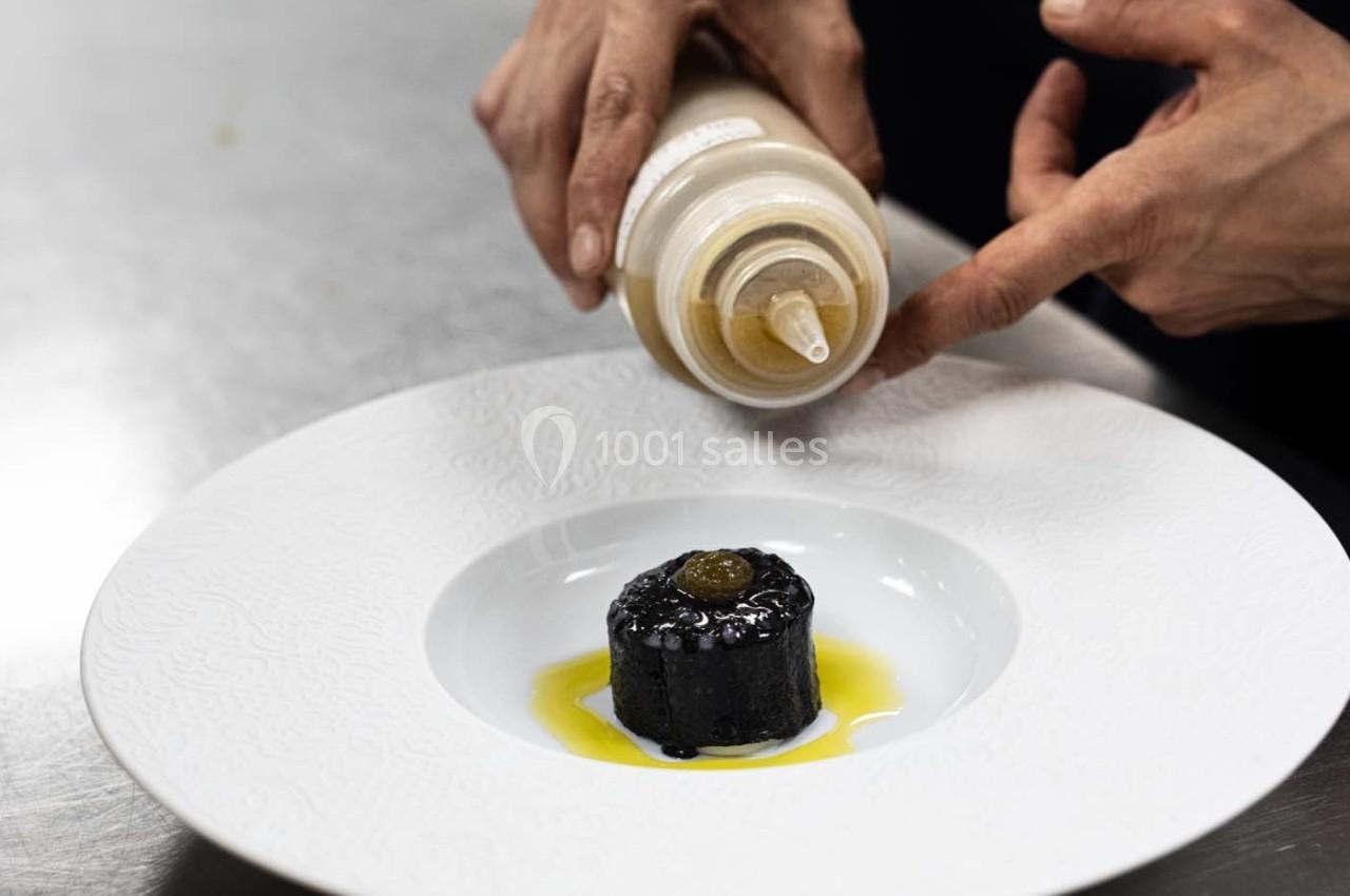 Une personne verse une sauce sur un plat dressé avec un cylindre noir surmonté d'une garniture dans une assiette blanche.