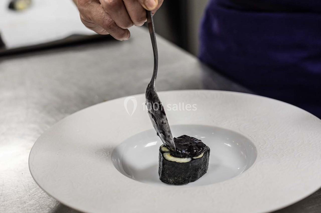 Un cuisinier dépose une sauce sombre sur un rouleau enrobé d'algue, placé au centre d'une assiette blanche.