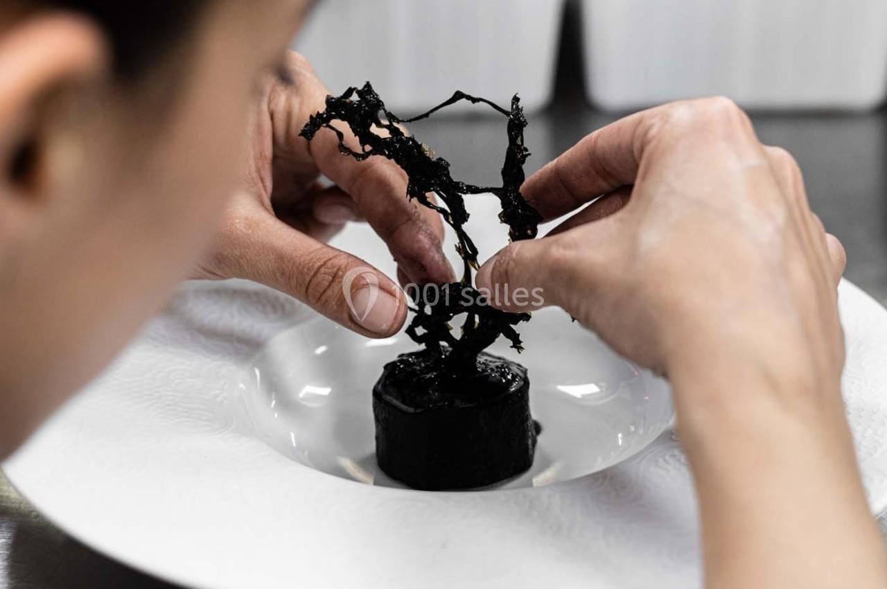 Une personne ajuste une décoration noire délicate sur un plat gastronomique dans une assiette blanche.
