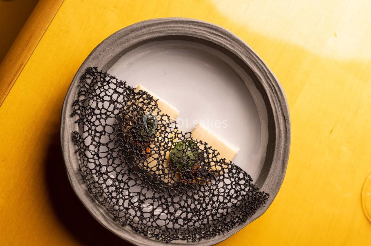 Assiette en céramique contenant une préparation gastronomique avec filet noir décoratif et morceaux de poisson.