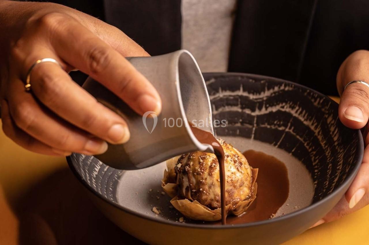 Une personne verse une sauce chocolatée sur un dessert composé d'une boule de glace et d'une base croustillante.