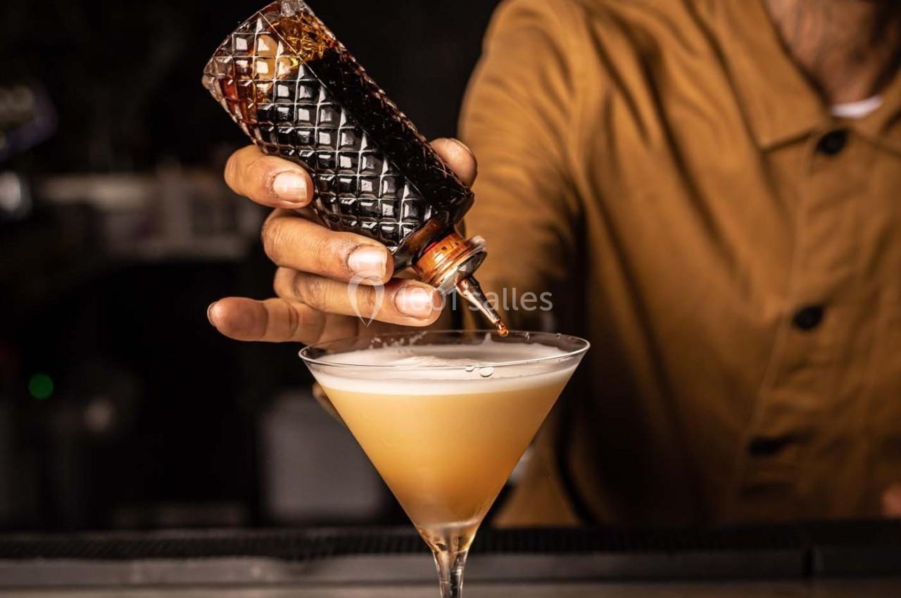 Un barman verse un liquide sombre dans un verre à cocktail rempli d'une boisson mousseuse.