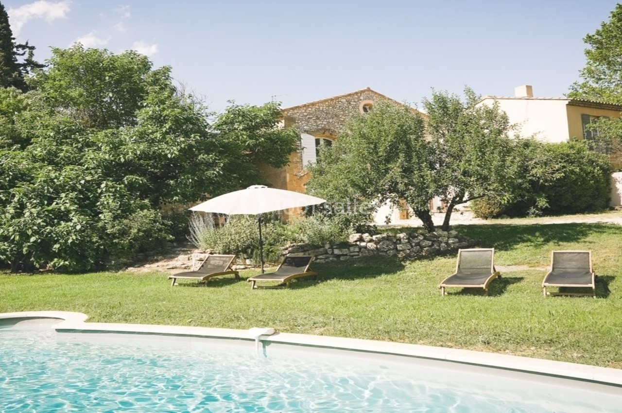 Jardin avec piscine, transats et parasol, entouré de verdure et d'une maison en pierre sous un ciel dégagé.