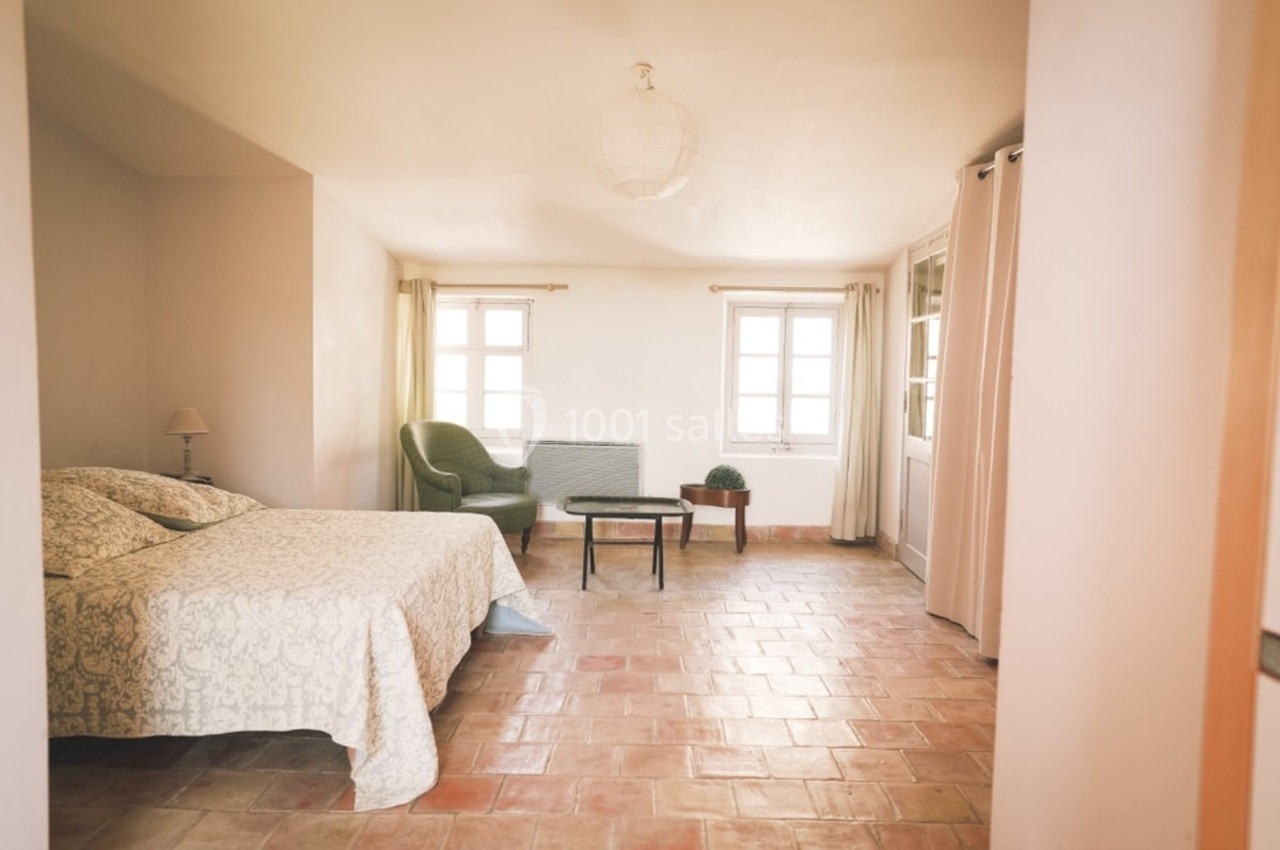 Chambre lumineuse avec lit double, fauteuil, table basse et sol en terre cuite, éclairée par deux fenêtres.