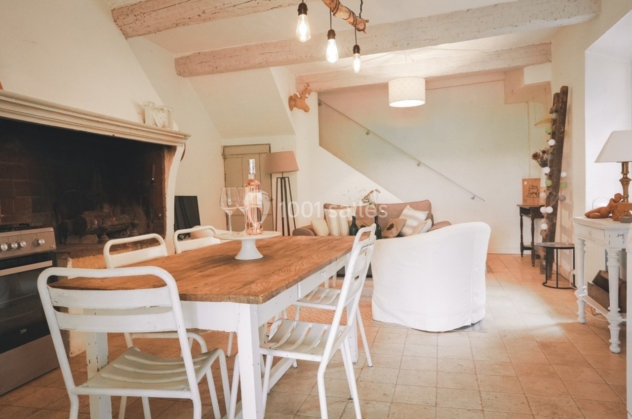 Salle à manger rustique avec table en bois, chaises blanches, cheminée et salon cosy en arrière-plan.