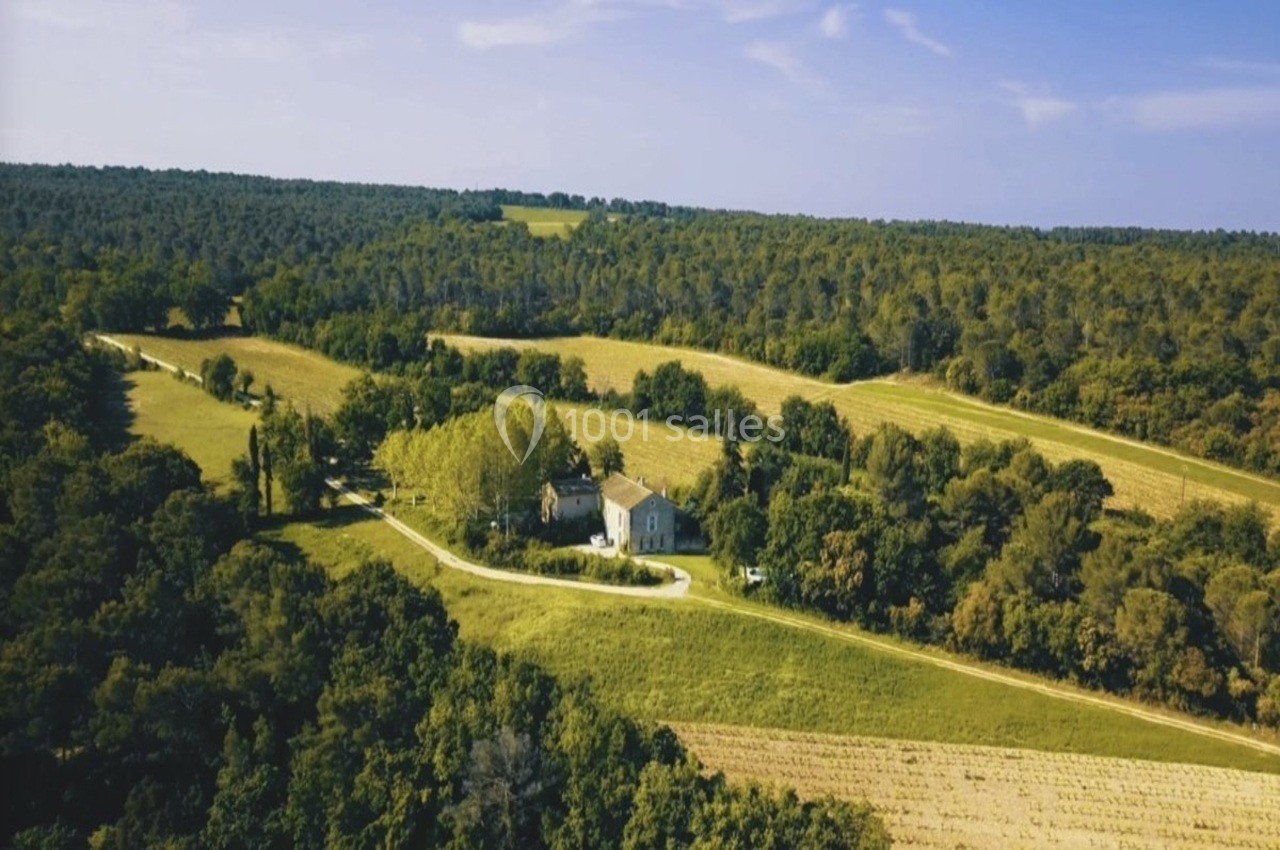 Vue aérienne d'une maison entourée de champs et de forêts sous un ciel bleu dégagé.