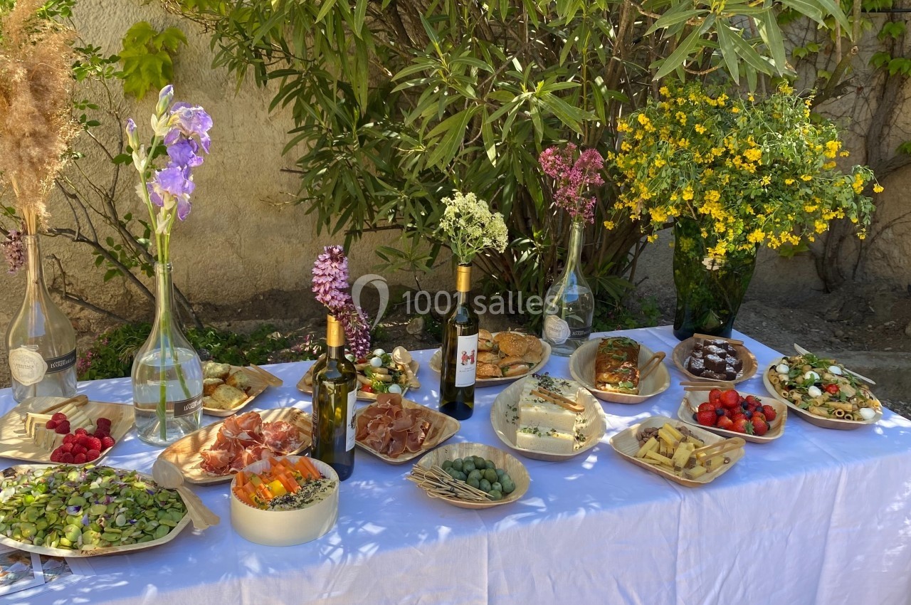 Table dressée en extérieur avec des plats variés, des fleurs en vase, des bouteilles de vin et un décor végétal en arrière…