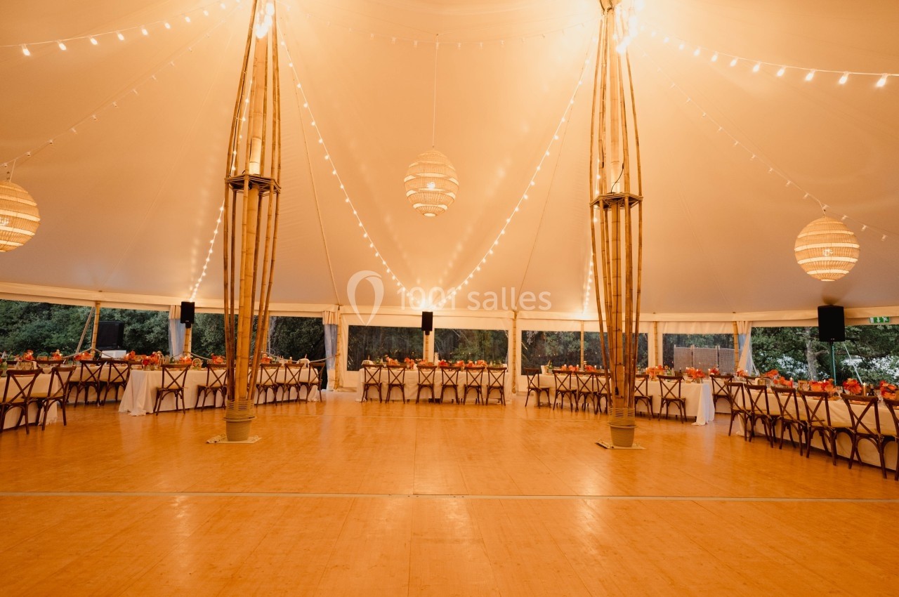 Salle de réception sous une tente éclairée avec des guirlandes lumineuses, tables décorées et espace central dégagé.