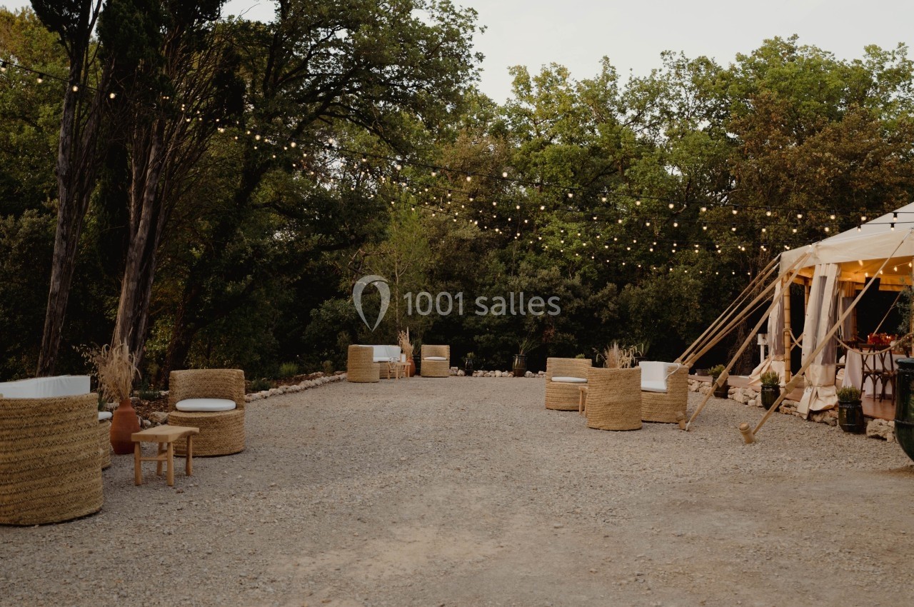 Espace extérieur décoré avec des fauteuils en osier, une tente blanche et des guirlandes lumineuses, entouré d'arbres.