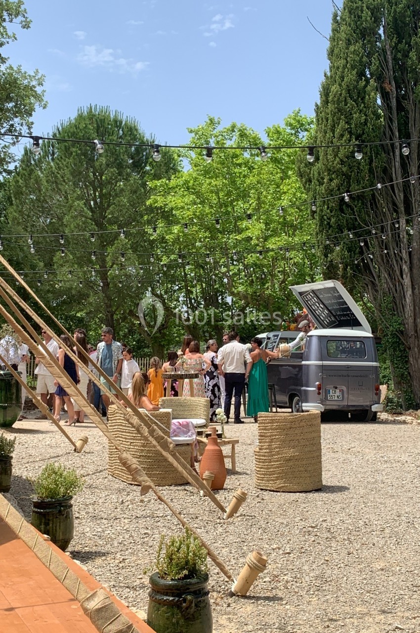 Un groupe de personnes rassemblé autour d'un camion-bar vintage dans un espace extérieur arboré et ensoleillé.