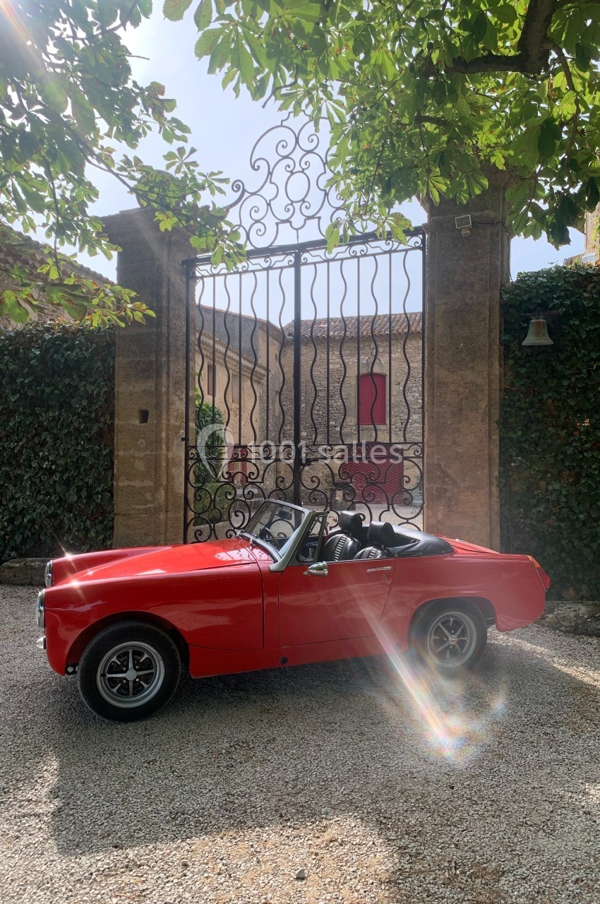 Voiture décapotable rouge garée devant un portail en fer forgé, entourée de végétation et baignée de lumière naturelle.