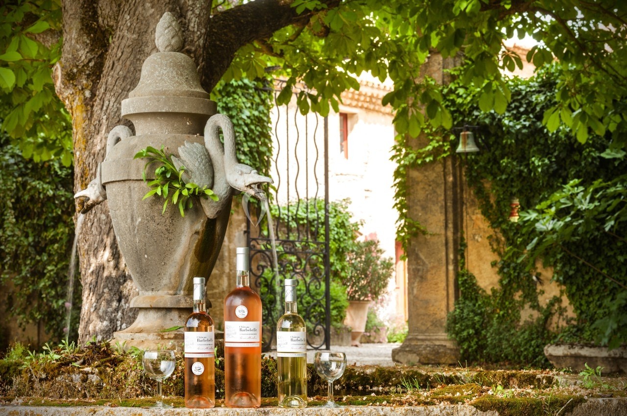 Trois bouteilles de vin et deux verres posés près d'une urne en pierre sous un arbre dans un jardin verdoyant.